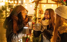 Drei Frauen stehen an einem Glühweinstand auf einem Weihnachtsmarkt in München und trinken heißen Glühwein