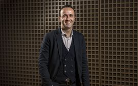 Ein lächelnder Mann im Anzug steht vor einer modernen Holzstruktur und wirkt professionell