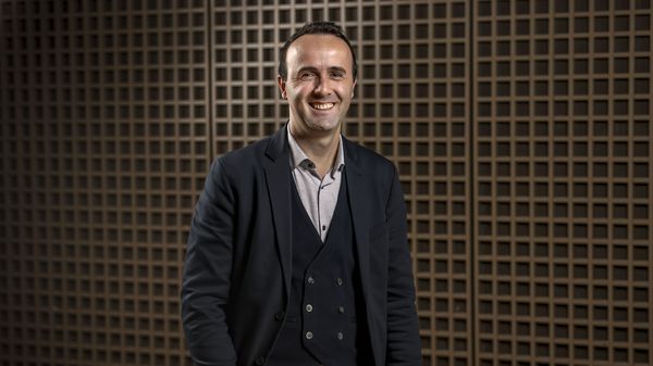 Ein Mann im Anzug steht lächelnd vor einer modernen Holzwand mit Gitterstruktur