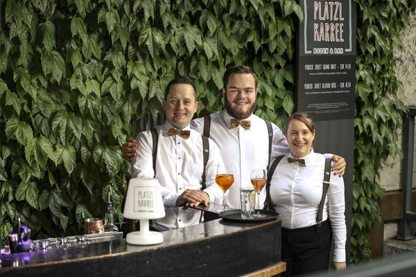 Drei Mitarbeitende des Platzl Karree stehen mit einem Serviertablett Arm in Arm vor dem Restaurant