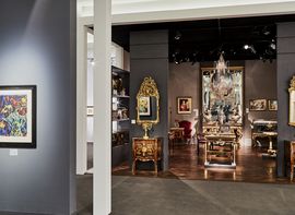 Ausstellungsraum in einer Kunstgalerie mit antiken Möbelstücken, vergoldeten Spiegeln, Kronleuchter und gerahmtem Bild an der grauen Wand im Vordergrund