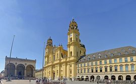 Die gelbe Theatinerkirche am Odeonsplatz in München vor einem blauen Himmel mit Passanten.