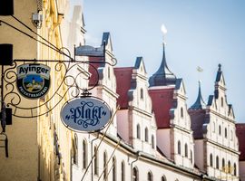 Ein Blick entlang Münchnener Häuserfassaden, an denen neben dem Logo der Brauerei Ayinger auch ein Logo des Platzl Hotels München hängt