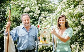 Zwei Personen mit Holunderprodukten vor blühenden Sträuchern im Garten
