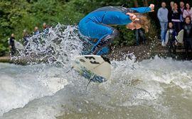 A surfer in blue wetsuit at the Eisbach wave in front of spectators.